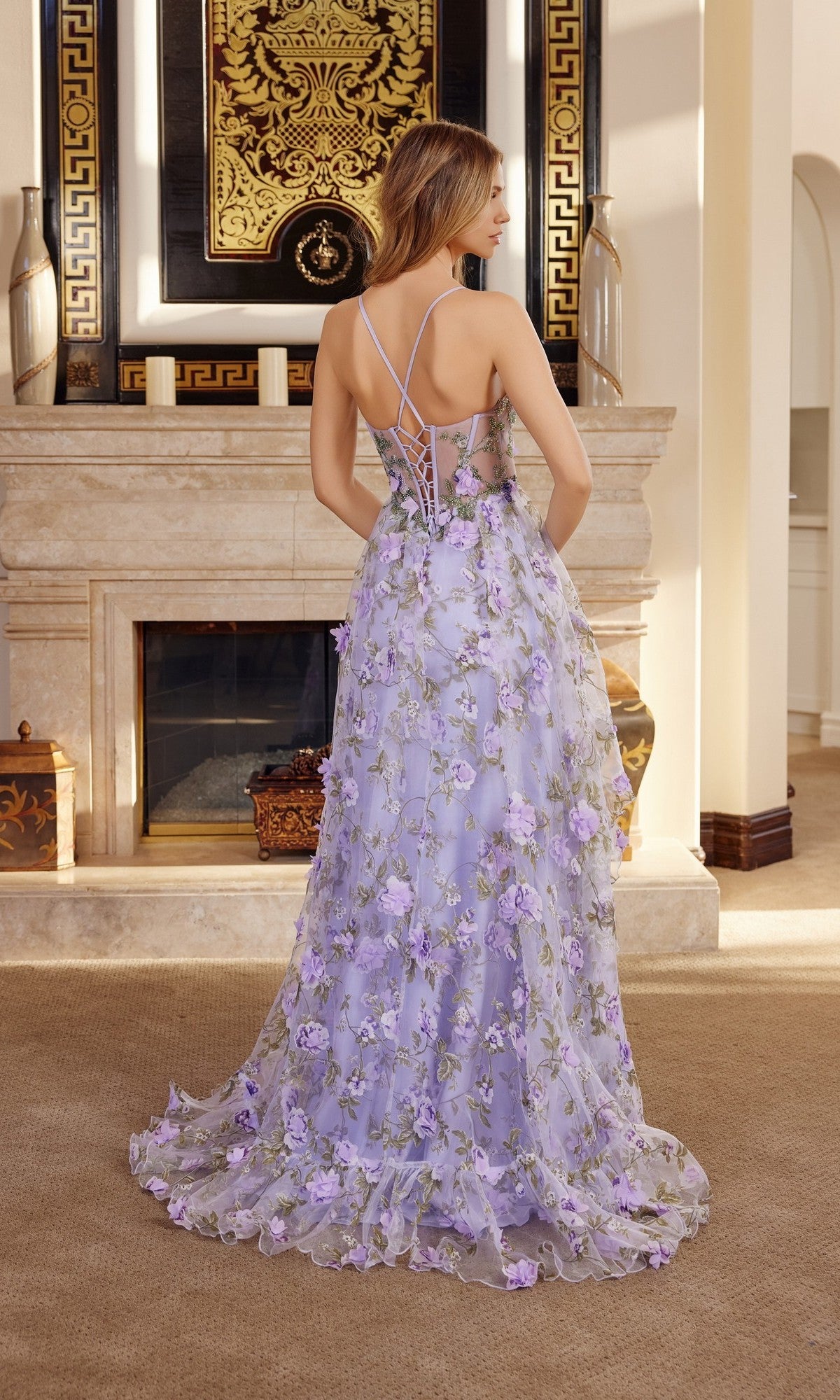 A woman in the Nox Anabel G1783 long prom dress, featuring a lavender floral print chiffon fabric, sheer back, and delicate straps, stands in front of a fireplace in an elegant room.